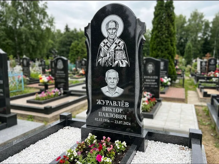 Памятник с гравировкой иконы: стела с образом в полный рост на обороте — гравировка образа Матроны Московской, низкие кусты спиреи в цвету, работа «Лебедева» в Калининграде